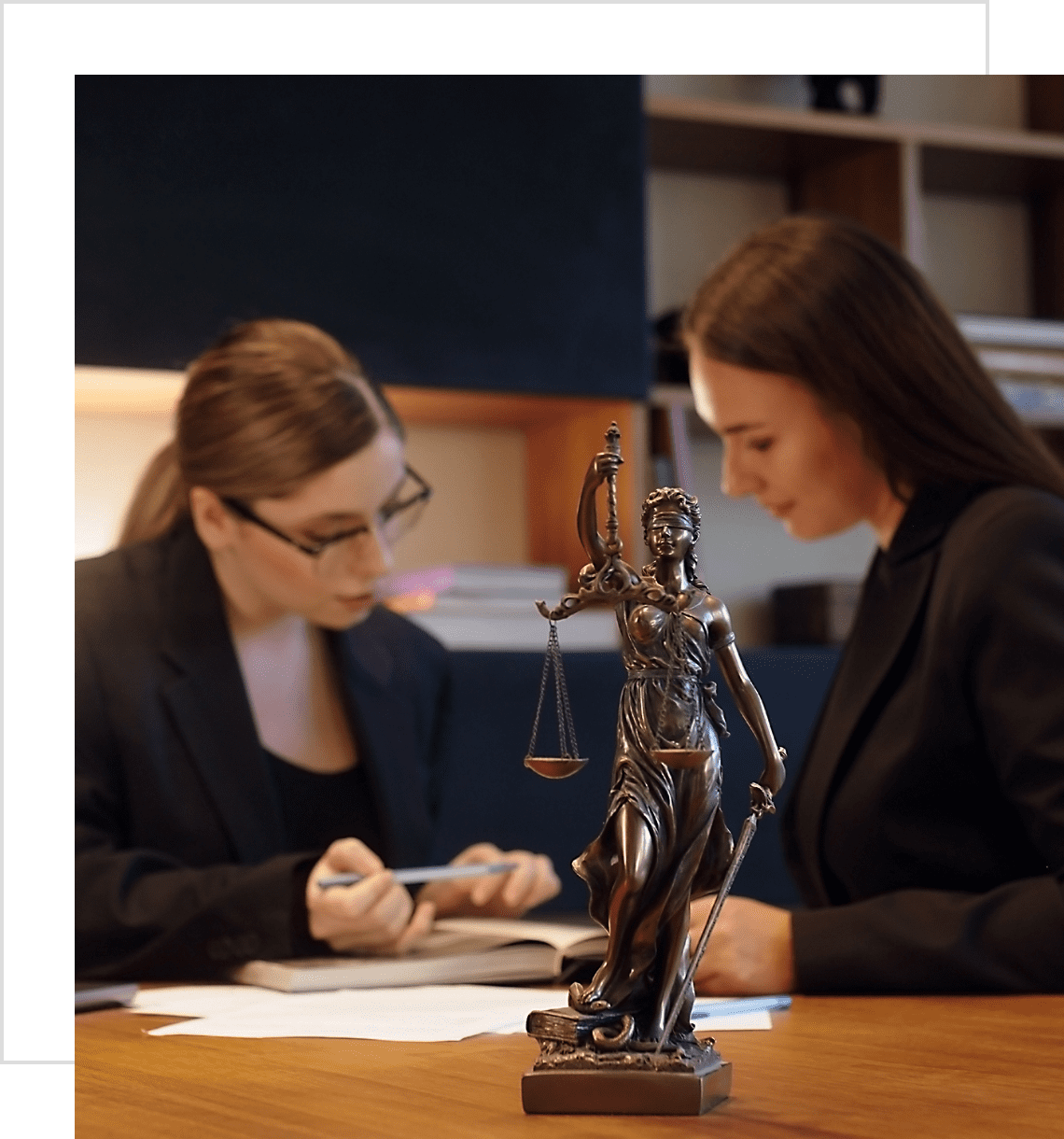 Two women discussing documents at a desk with a Lady Justice statue.
